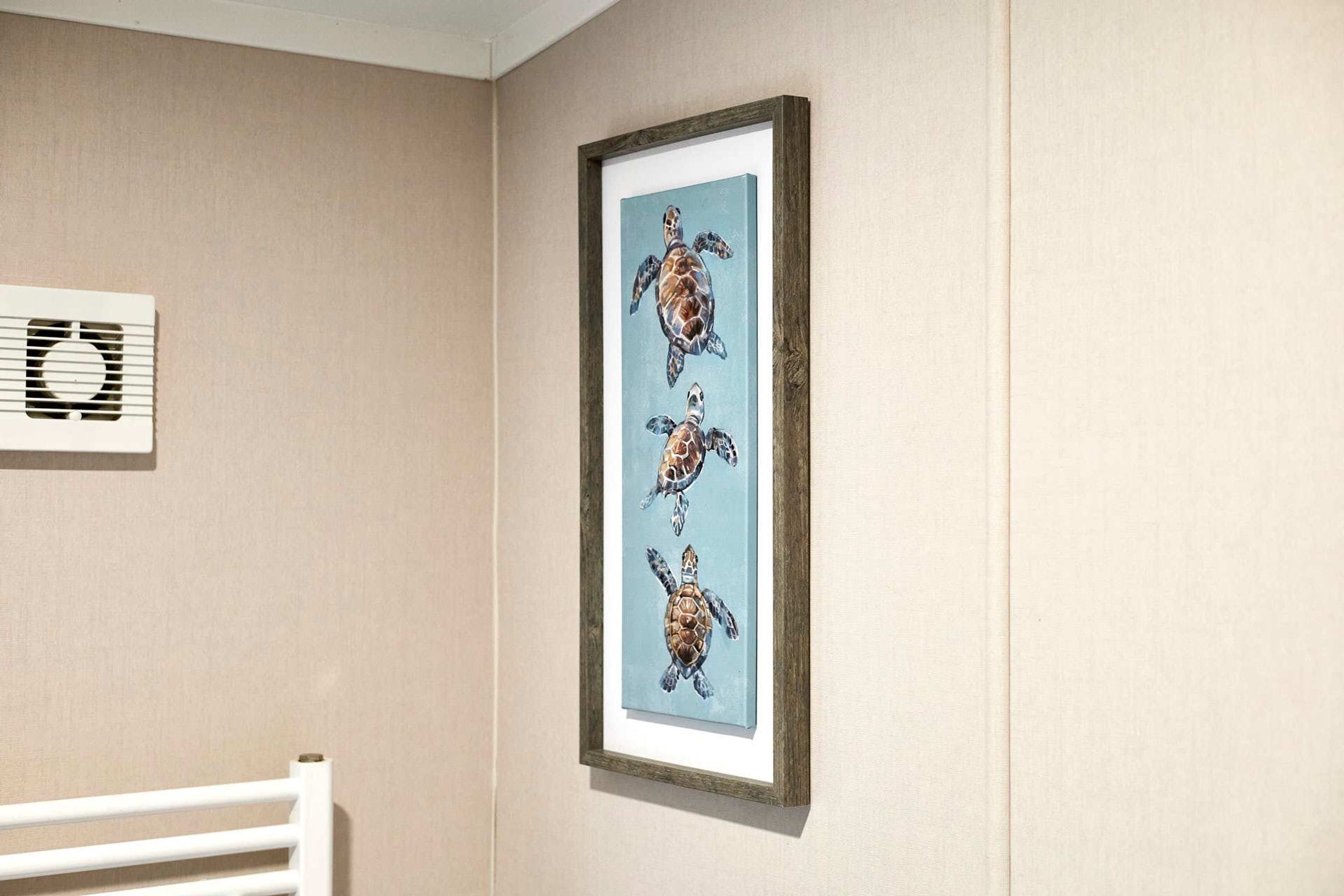 A framed artwork of three sea turtles on a light blue background, mounted on a beige wall.
