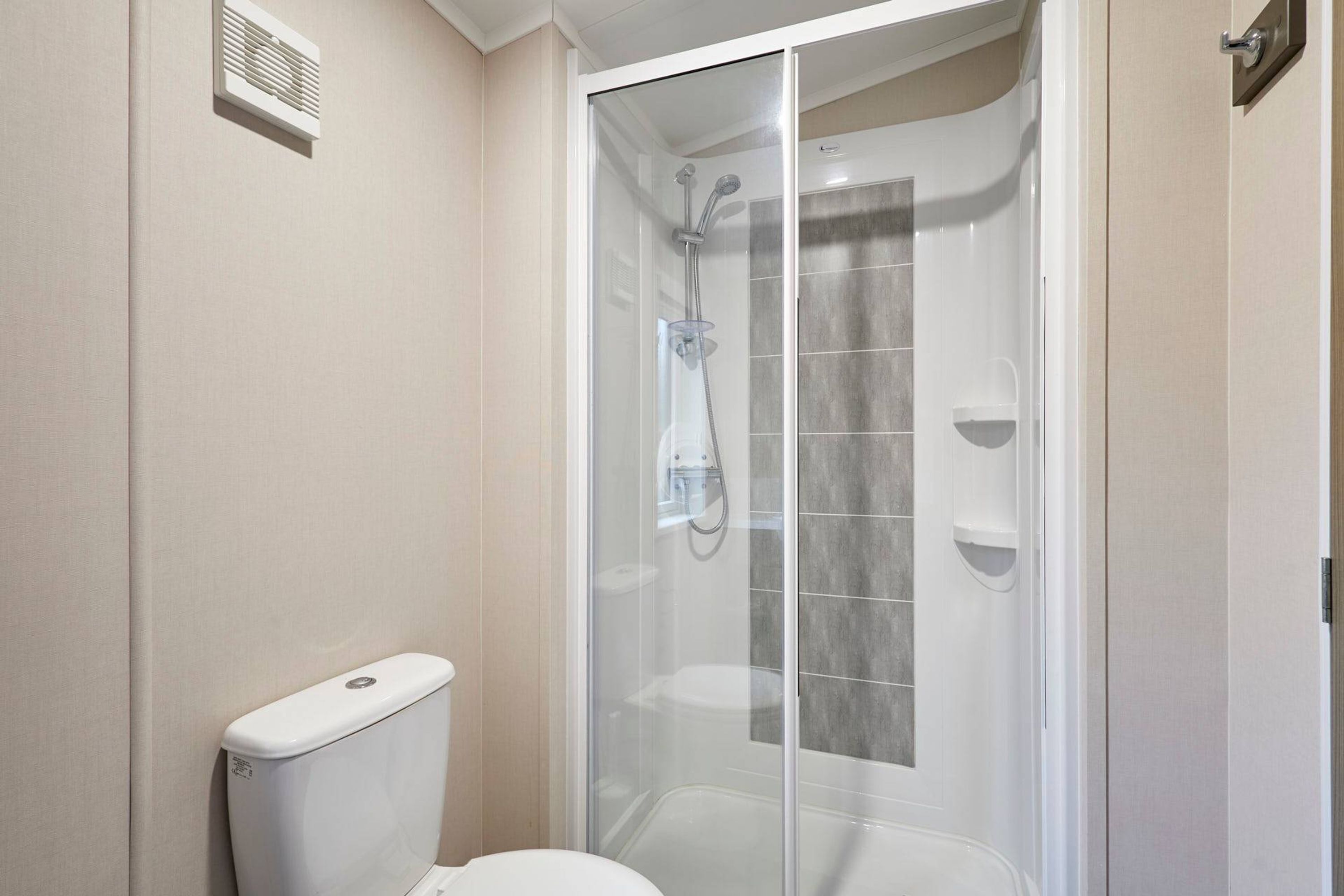 Baño con ducha de vidrio, inodoro y extractor de aire empotrado.