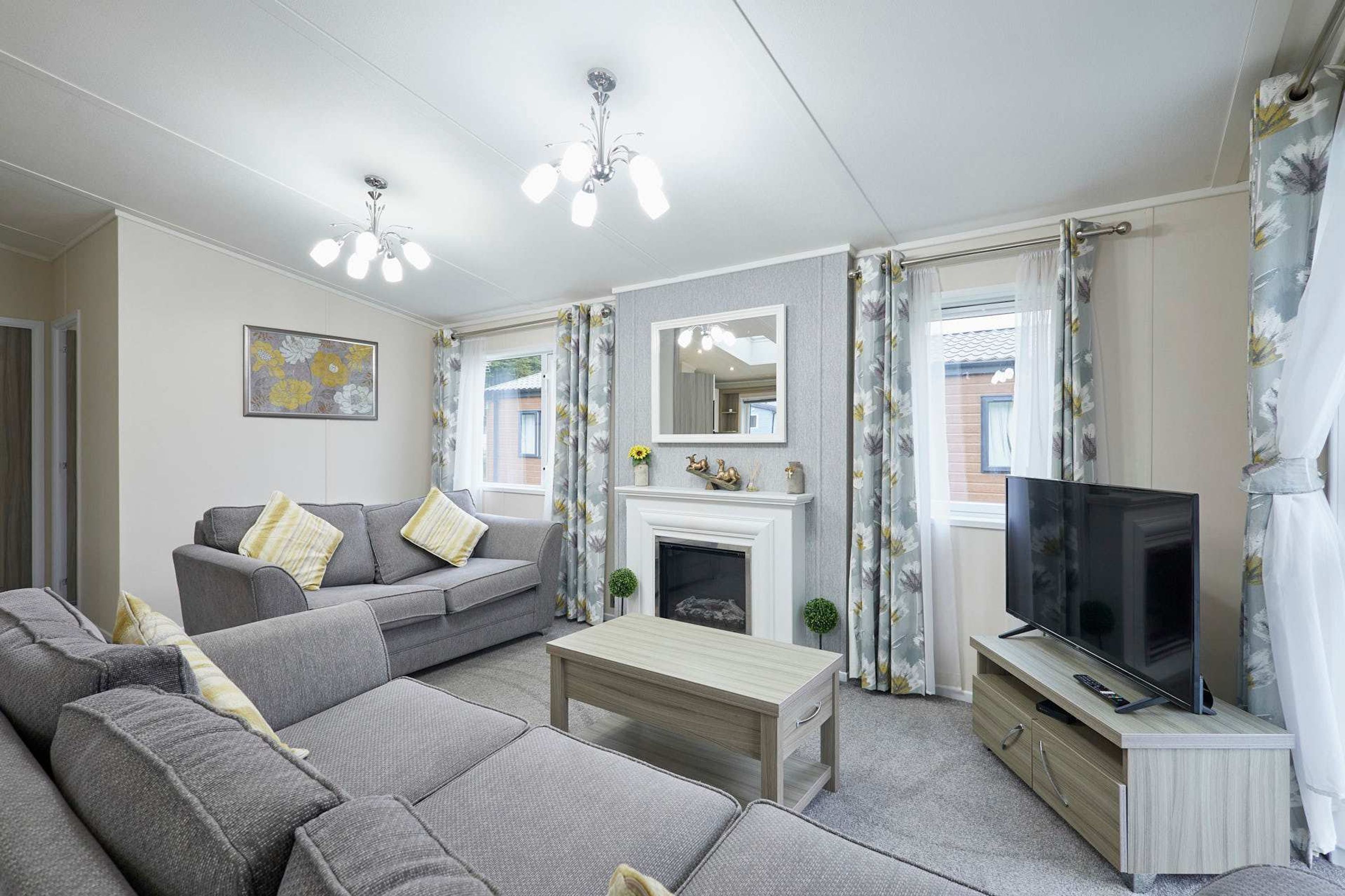 A modern living room with a grey sofa set, a wooden coffee table, a flat-screen TV on a wooden stand, a fireplace with a mirror above it, and floral curtains.