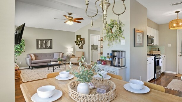 A modern living room with a sofa, coffee table, and dining table set with white dishes and a centerpiece. A kitchen with appliances and a ceiling fan is visible in the background. - Howdy Hang Out - Cozy, Cute And Comfortable House! 2 Bedroom Home (Bryan)