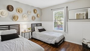 A bedroom with two beds, a bedside table with a lamp, a wall-mounted mirror, and a window with blinds. - Howdy Hang Out - Cozy, Cute And Comfortable House! 2 Bedroom Home (Bryan)