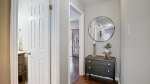 A hallway with a white door, a dark dresser with drawers, a round mirror, and a small plant on a wooden stand. - Howdy Hang Out - Cozy, Cute And Comfortable House! 2 Bedroom Home (Bryan)