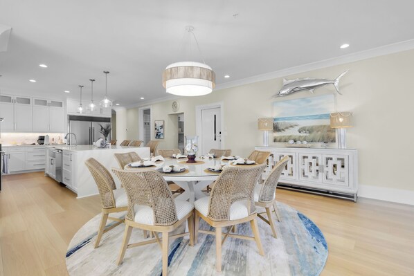 A modern kitchen with white cabinetry, a central island, and a dining area with wicker chairs and a round table. - Mar-Linn - Penthouse - Sleeps 12 at Big Rock Landing (Morehead City)