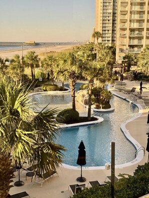 Ein Poolbereich eines Resorts mit Palmen, Liegestühlen und Blick auf Strand und Meer.