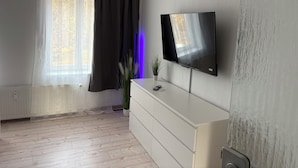 Una habitación moderna con un televisor de pantalla plana grande montado en la pared, un tocador blanco, una planta en maceta y una ventana con cortinas translúcidas.