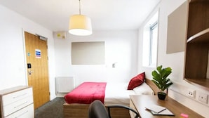 Una habitación pequeña y luminosa con una cama, un escritorio, una silla, un radiador, una puerta de madera y una ventana.