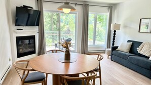 Un salon avec une table ronde en bois, quatre chaises, un canapé bleu foncé, un téléviseur fixé au mur et une cheminée.