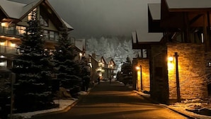 Une rue enneigée bordée de bâtiments des deux côtés, illuminée par des lumières chaleureuses, avec une chaîne de montagnes en arrière-plan.