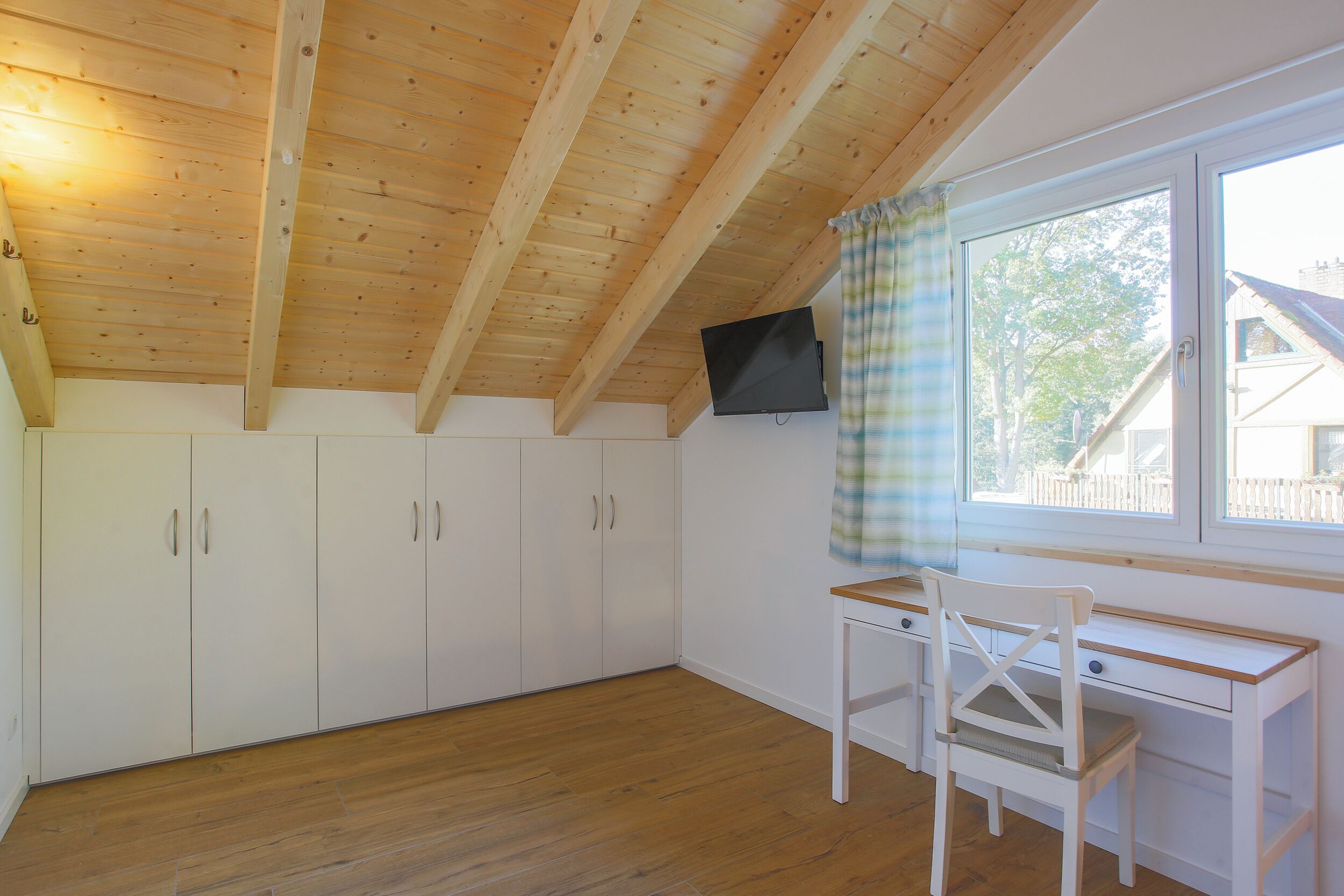 Una habitación con vigas de madera en el techo, armarios blancos, un escritorio y una silla.