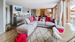 Un salon moderne avec un canapé d’angle, un petit pouf et un fauteuil blanc à poils longs. On aperçoit à l’arrière-plan une cuisine avec des meubles en bois et un coin repas comprenant une table et des chaises.