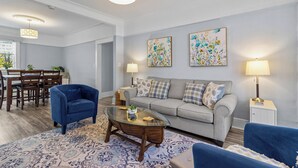 Ein Wohnzimmer mit einer Couch, Sesseln, einem Glastisch und einem Essbereich mit Tisch und Stühlen.
