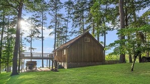 En stuga byggd av trä med en veranda och en båt förtöjd utanför, omgiven av träd och en klar himmel.