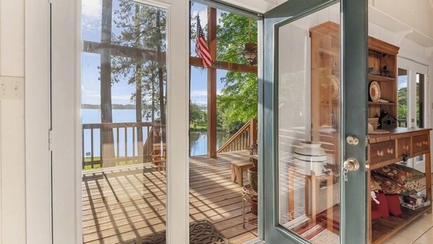 En träaltan med utsikt över en sjö och träd, en glasdörr som leder in till ett rum med ett träskåp och en liten modell av ett hus.