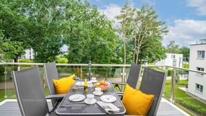 Un balcón con una mesa lista para comer, rodeado de sillas con cojines y vistas a vegetación y edificios.