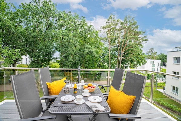 A balcony with a table set for dining, surrounded by chairs with cushions, and a view of greenery and buildings. - HaffSeits Zirchow - a modern vacation home nestled between nature and the lagoon (Zirchow)
