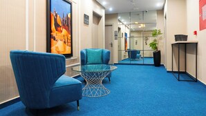 Un hall de hotel con alfombra azul, dos sillones azules, una mesa de centro de vidrio, un cuadro grande enmarcados de un paisaje desértico y una planta en maceta.