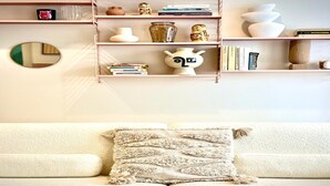 Una sala de estar con un sofá beige, un cojín decorativo y una estantería con diversos objetos.
