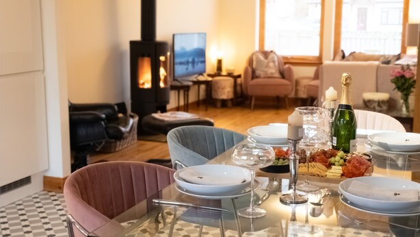 A dining area with a glass table set for a meal, a fireplace, and a cozy living room in the background. - Casa Amor - Aviemore (Aviemore)