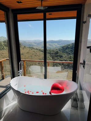 A modern bathroom with a large bathtub, a view of rolling hills, and a wooden ceiling.