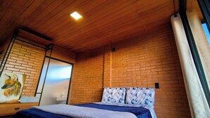 A bedroom with a brick wall, a wooden ceiling, a bed with patterned bedding, and a painting of a fox. - Ibitipoca Castle (Conceição da Ibitipoca)