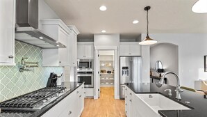 A modern kitchen with white cabinetry, a black countertop, stainless steel appliances, and a wooden floor. - Modern Family Home | Sauna, Fire Pit, Swings & Access to Pools & Parks (Kuna)