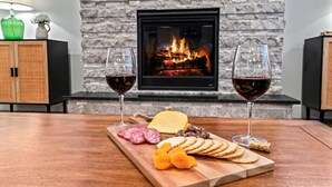 A cozy room with a stone fireplace, a wooden table with a cheese board, two glasses of red wine, and a lamp. - Modern Family Home | Sauna, Fire Pit, Swings & Access to Pools & Parks (Kuna)