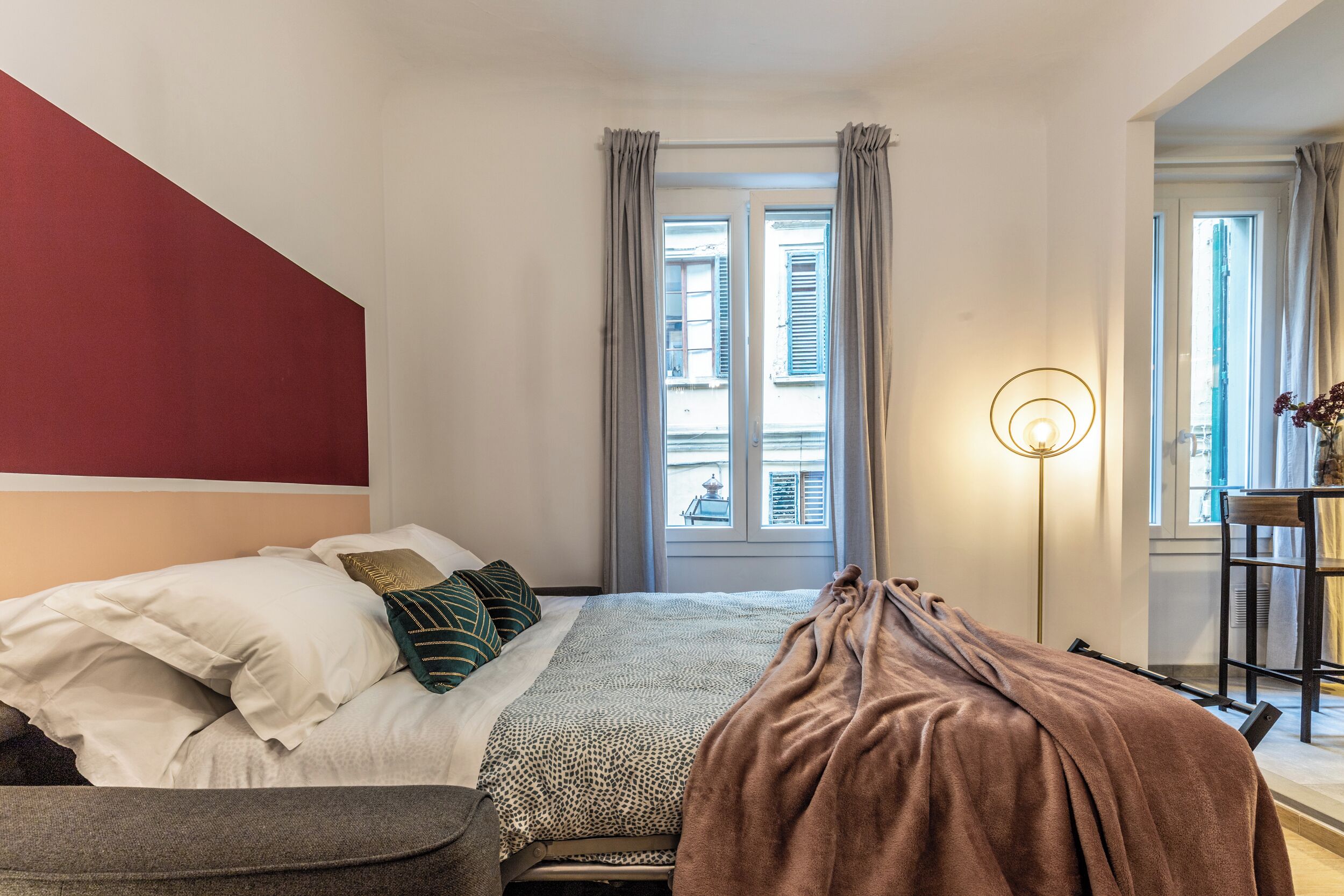 Et soveværelse med seng, natbord, lampe og udsigt til gaden gennem et vindue.