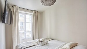 A neatly made bed with a folded towel, a window with curtains, and a hanging light fixture. - New Apartment Center of Paris Le Marais (Paris)