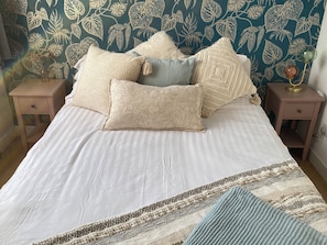 Ein Bett mit weißer Bettwäsche und mehreren Kissen, ein Nachttisch mit einer Lampe und eine Wand mit einem Blattmuster.