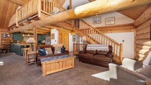 Ein geräumiges Blockhaus-Interieur mit hoher Decke, einer Holztreppe und einem gemütlichen Wohnbereich mit Couch, Couchtisch und Kamin.