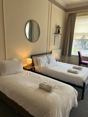 A hotel room with two single beds, a round mirror, a bedside table with a lamp, a window with curtains, and a view of the street outside. - Edinburgh Travel Guest House (Edinburgh)