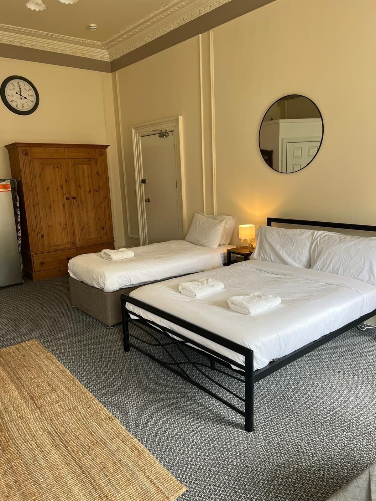 A hotel room with two single beds, a wooden wardrobe, a round mirror, and a clock on the wall.
