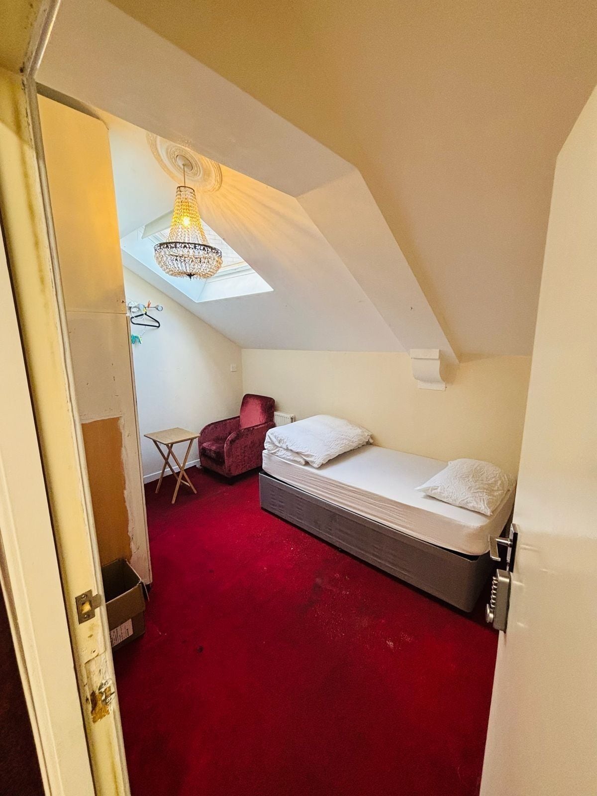 Een kleine, smalle kamer met een rood tapijt, een enkel bed, een rode stoel en een tafeltje.
