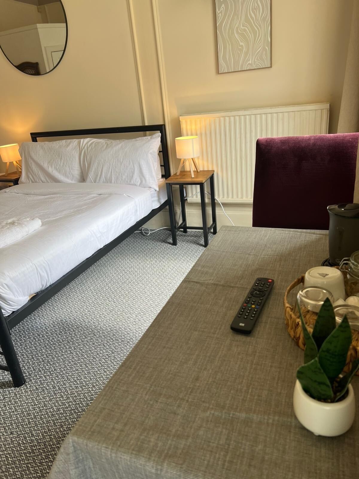 A neatly arranged hotel room with a bed, bedside lamp, chair, remote control, and a small table with a plant and candles.