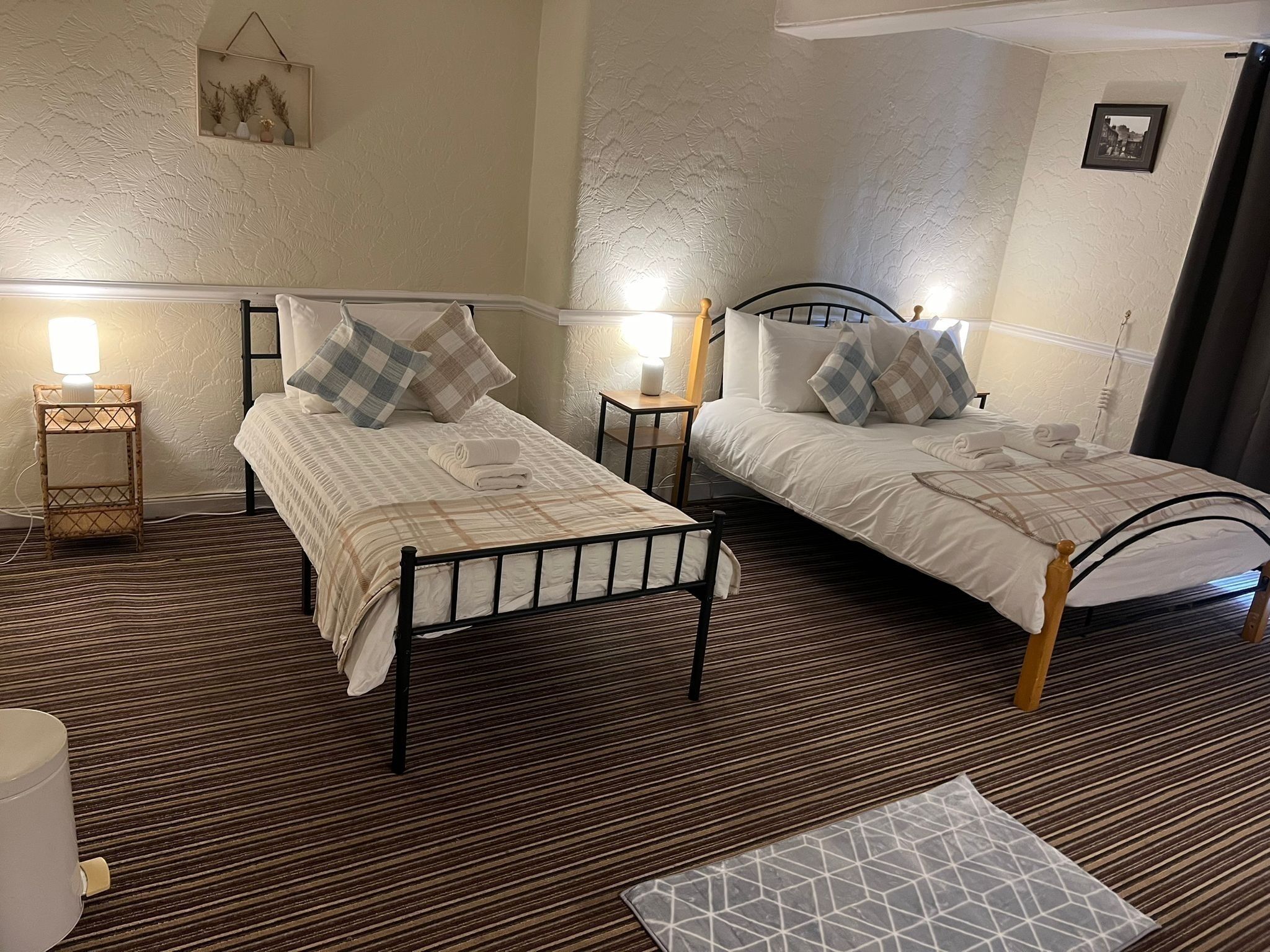 A room with two beds, each with a headboard, bedside tables, and lamps. The room has a patterned carpet and a small rug on the floor.