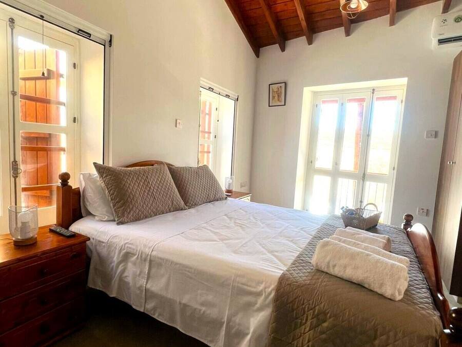 Un dormitorio con techo de madera, una cama con ropa blanca, una mesita de noche con lámpara y una ventana con cortinas.