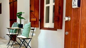 Una habitación con puerta de madera, ventana con contraventanas, una mesita con sillas y una planta en maceta.