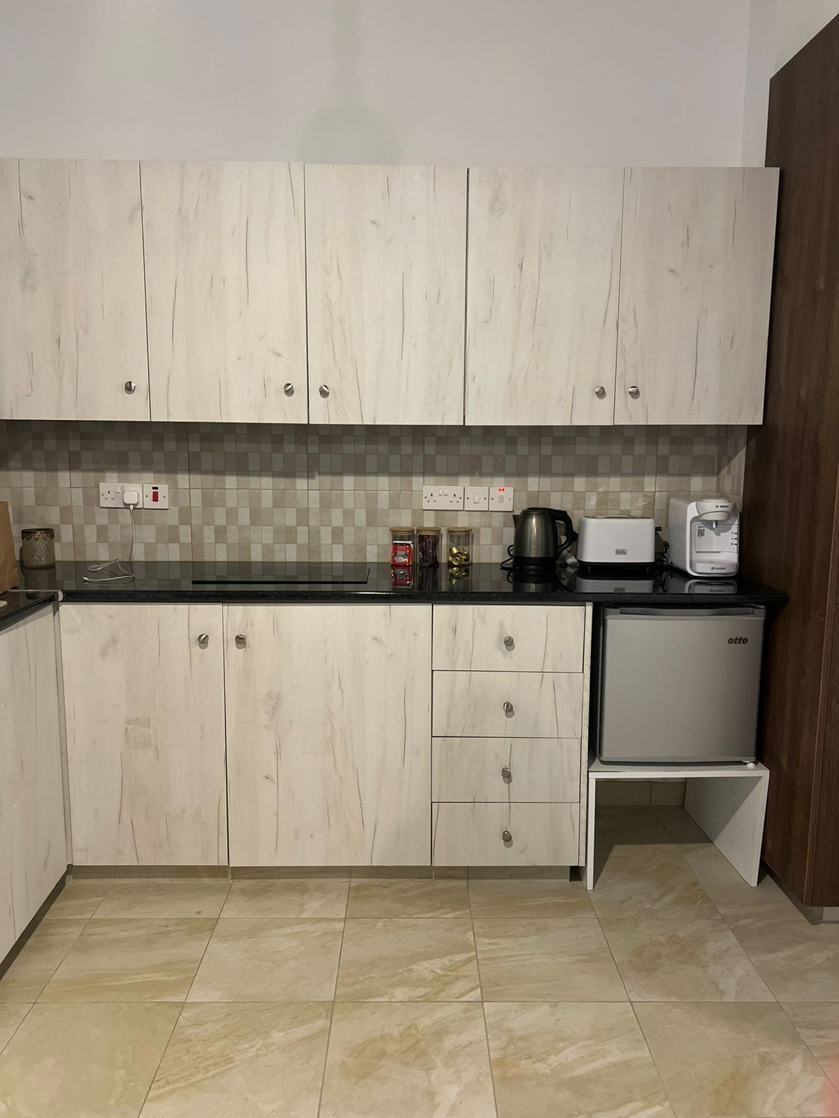 Una cocina con armarios de madera clara, salpicadero azulejado, cafetera, tostadora y refrigerador pequeño.