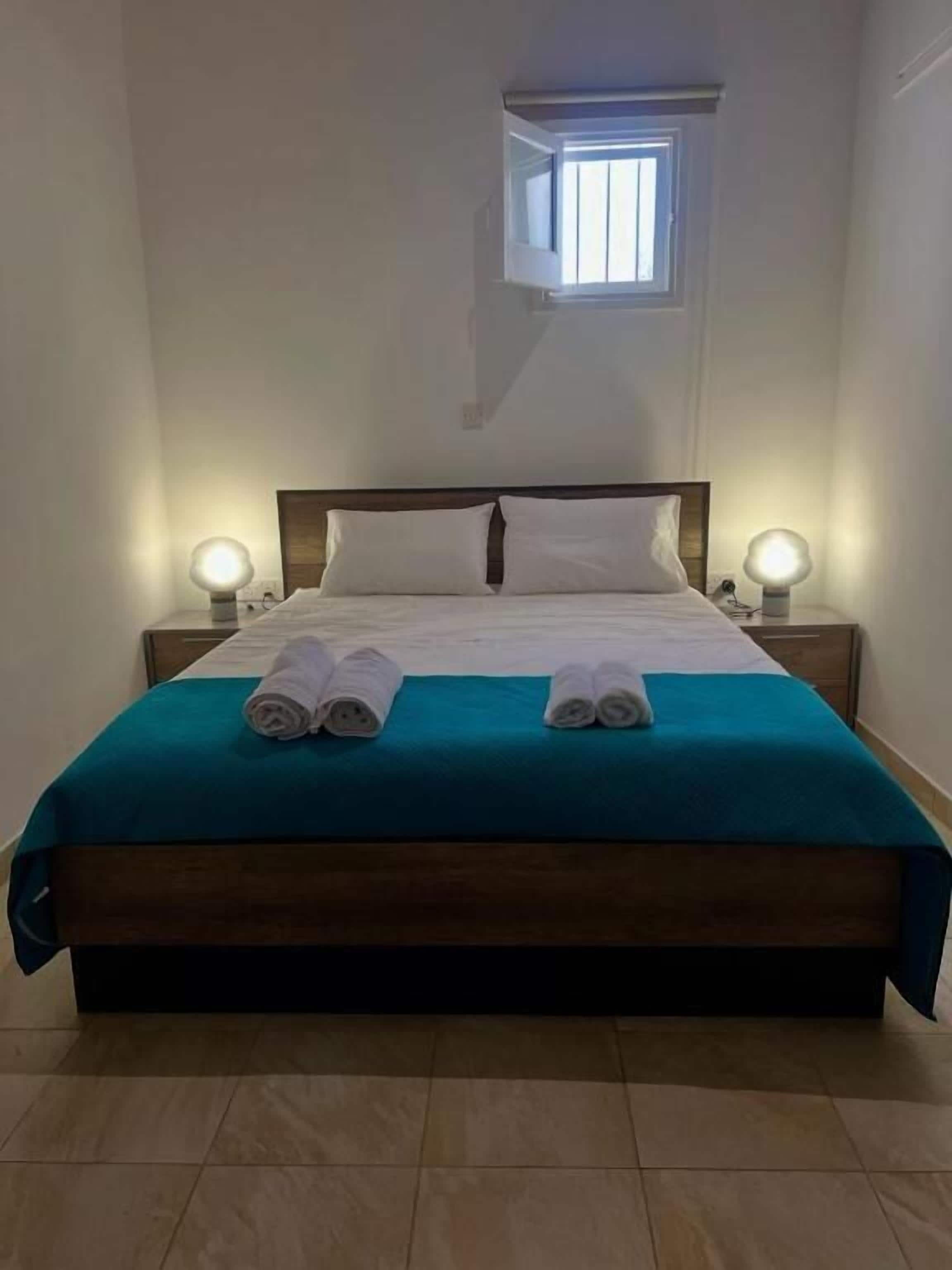 Una cama bien hecha con dos toallas enrolladas, un cabecero de madera y dos lámparas de noche.