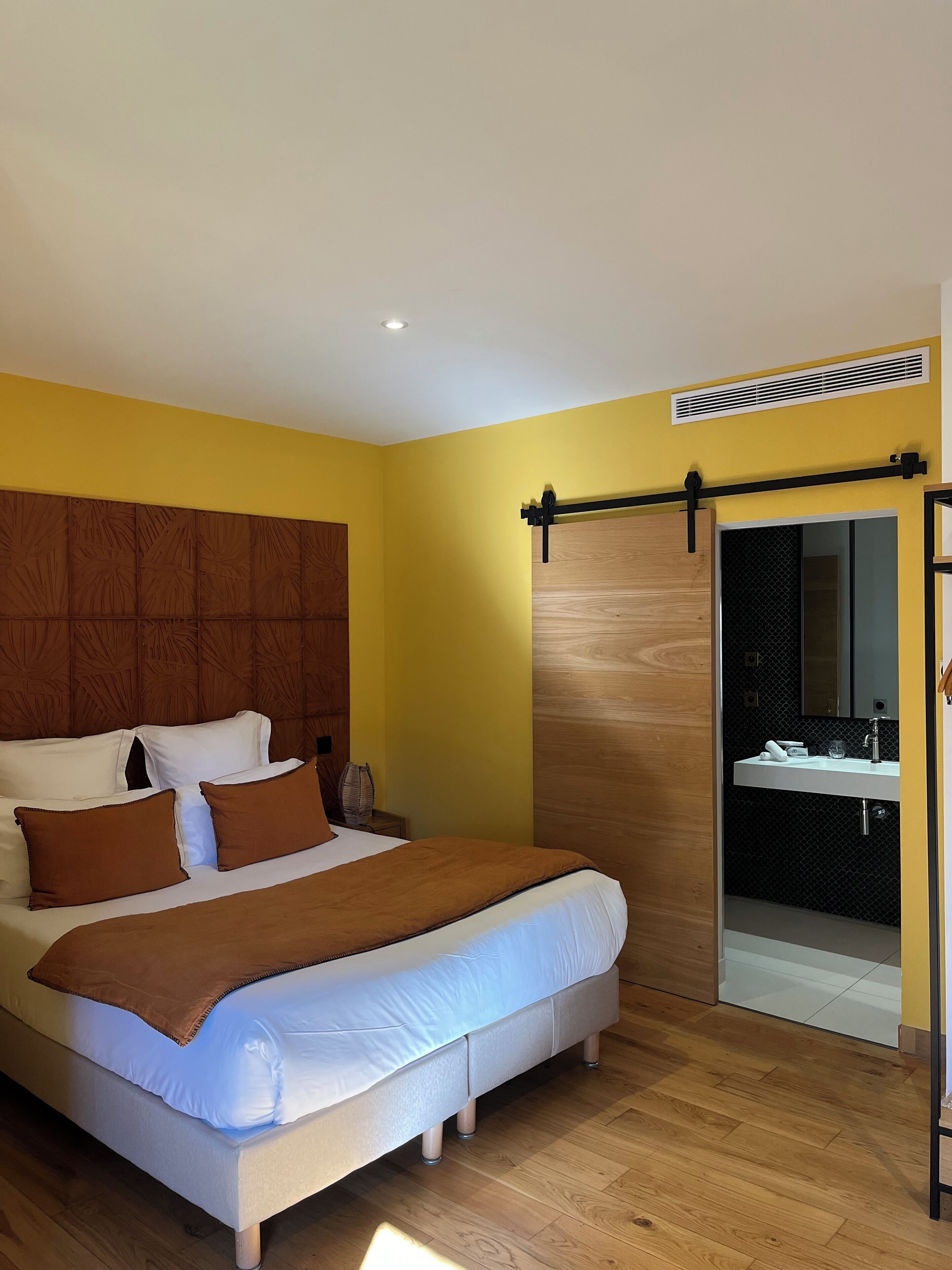 Un dormitorio con cama, cabecera de madera y puerta corrediza que da a un baño con lavabo y espejo.