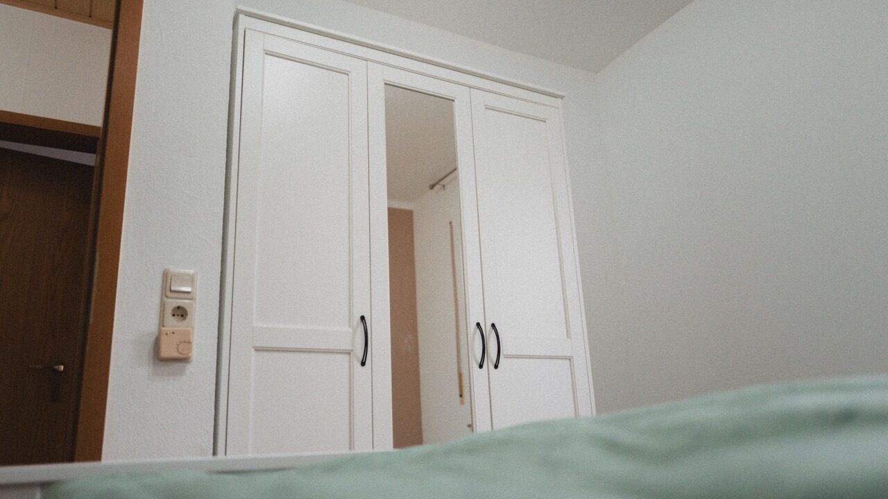 A room with a white wardrobe, a wooden door, and a light switch on the wall.