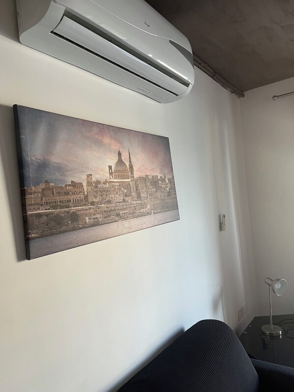 A room with a mounted air conditioner, a framed cityscape painting, a dark-colored sofa, and a small table with a lamp. - Charming seafront 2-bedroom apartment in lovely St. Julian's with AC (St. Julian's)