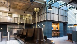 Uma área de espera moderna com um espaço de estar central, bar e teto alto com vigas aparentes.