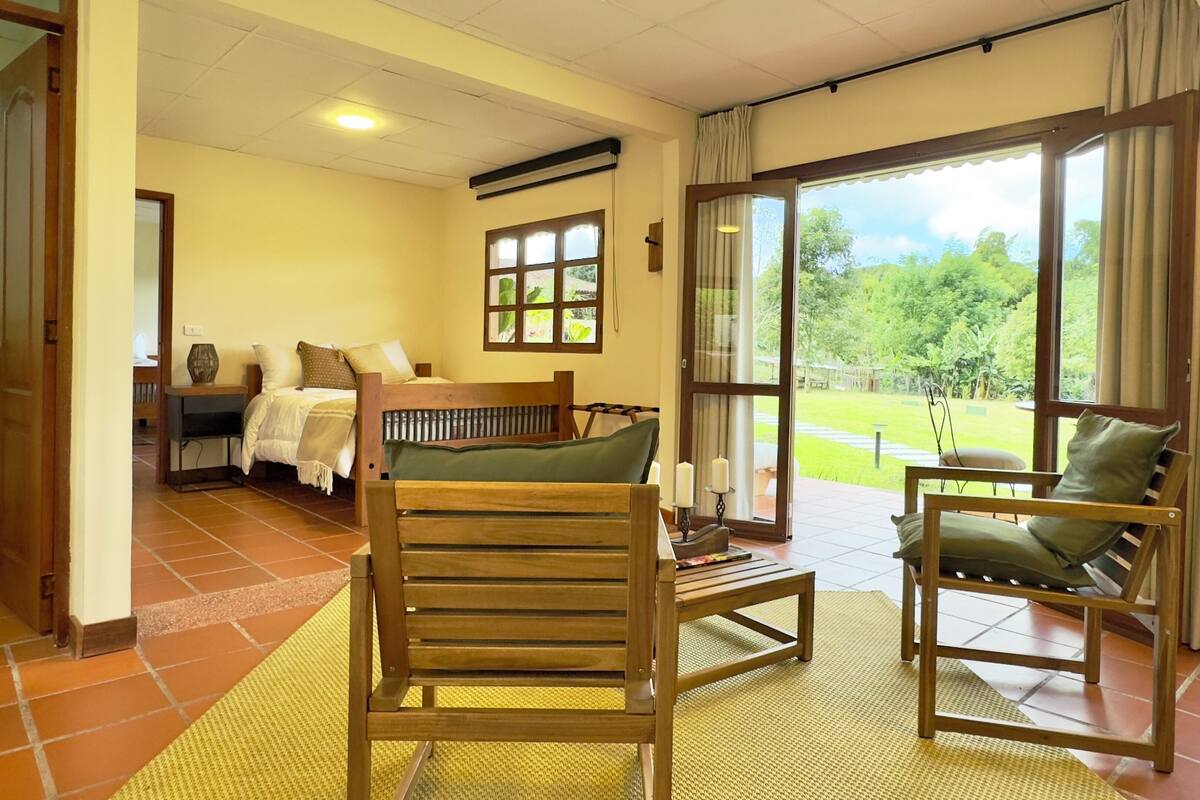 Una habitación con una cama, una silla de madera, una mesa y vistas a un jardín a través de ventanas amplias.