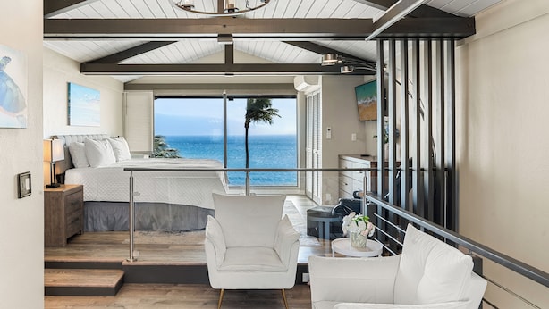 Een slaapkamer met een groot raam met uitzicht op de oceaan, een bed met witte lakens, een nachtkastje en een stoel.
