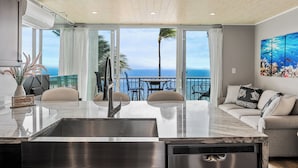 Een moderne keuken met een groot eiland, een eethoek met stoelen en een tafel, en een balkon met uitzicht op de oceaan.