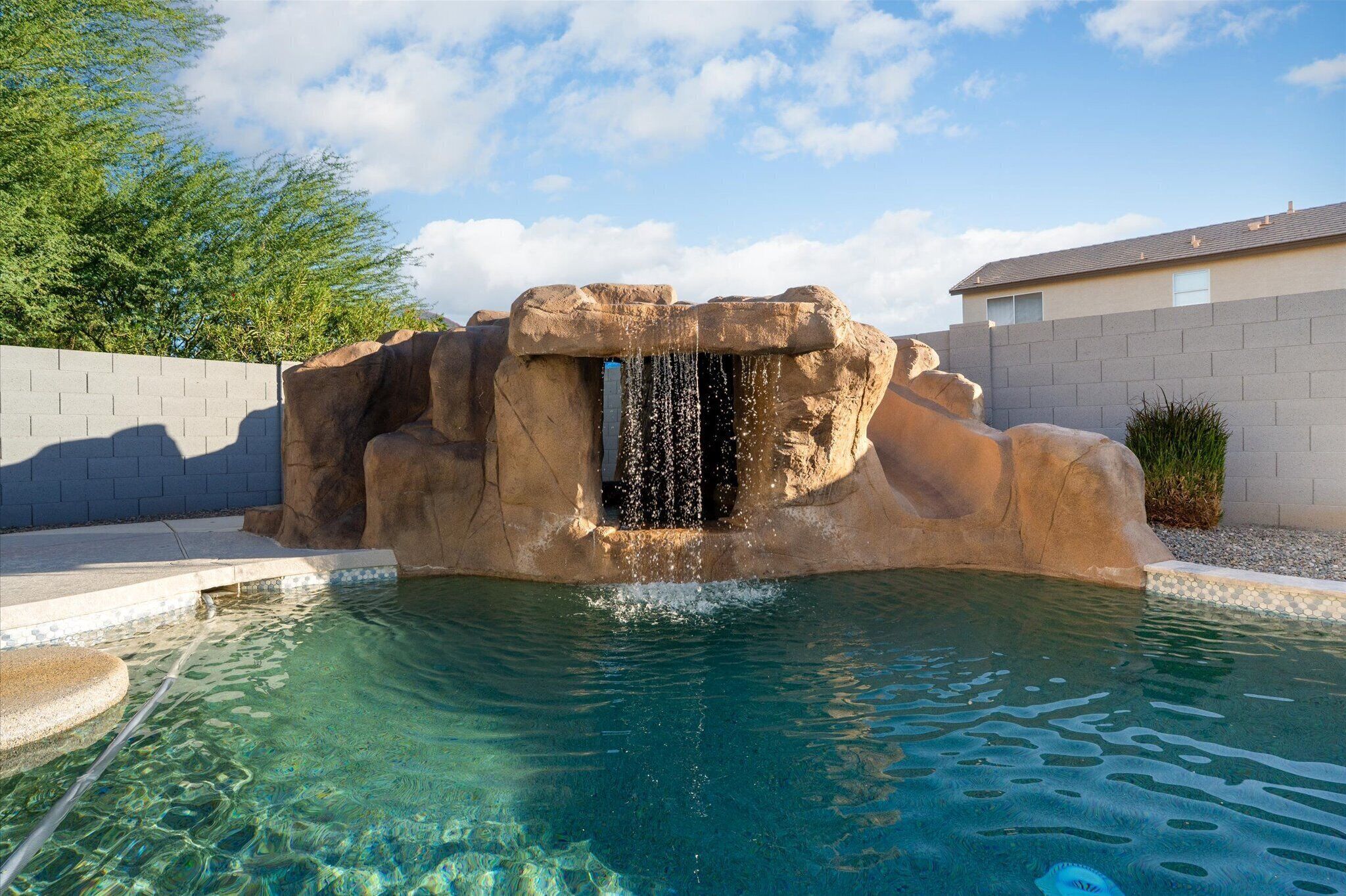 A clear pool with a rock waterfall feature, surrounded by a concrete wall and a small garden area.