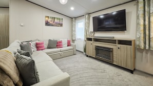 A living room with a sectional sofa, a flat-screen TV, a fireplace, and a large window with curtains. - Host & Stay | Happy Day's Lodge (Pocklington)