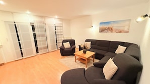 Een woonkamer met een zwarte bank, een houten salontafel en een stoel. Er is een schuifdeurkast en een schilderij aan de muur.
