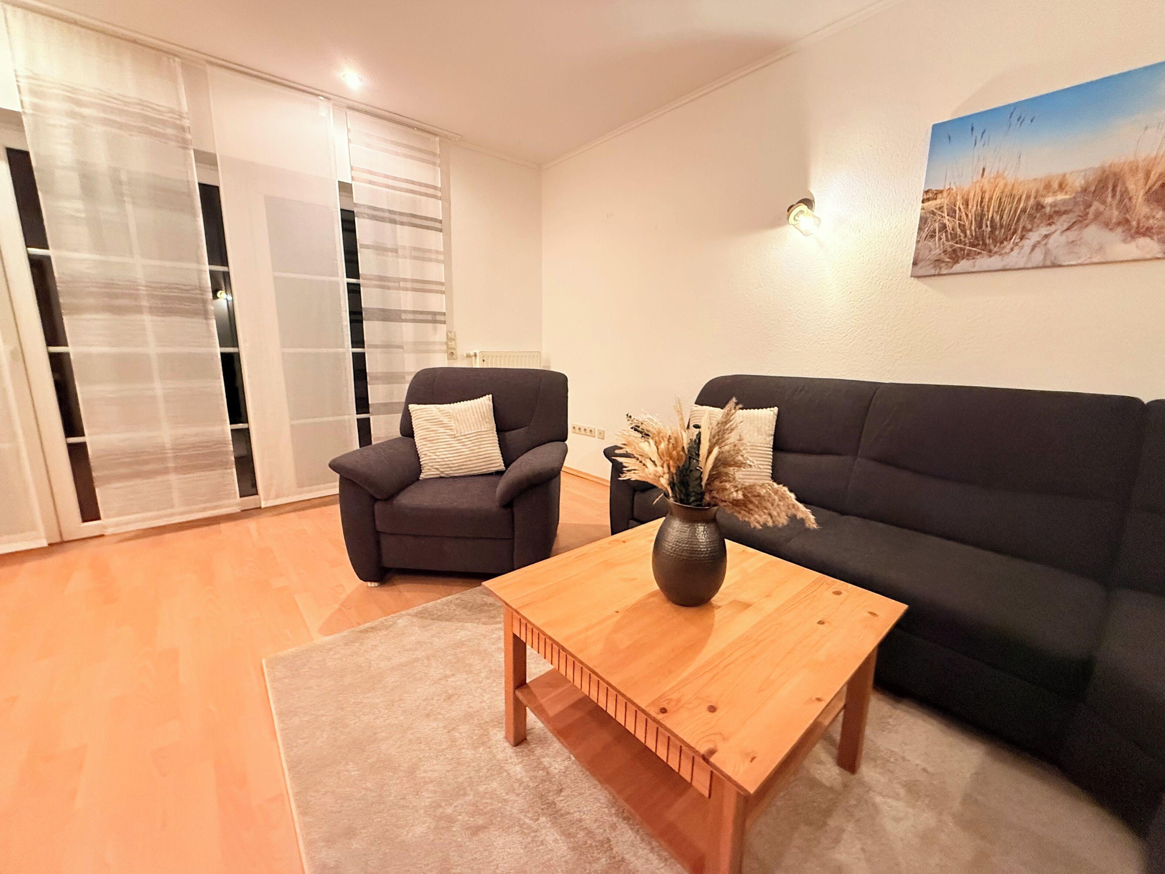 A living room with a dark grey sofa, a wooden coffee table with a vase of dried plants, and a black armchair with a striped pillow.